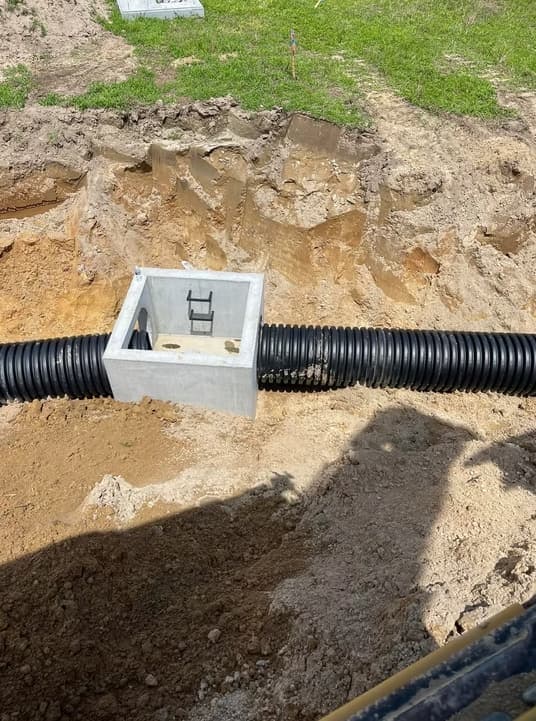 A precast catch basin connected to corrugated drainage pipe in a freshly dug trench.