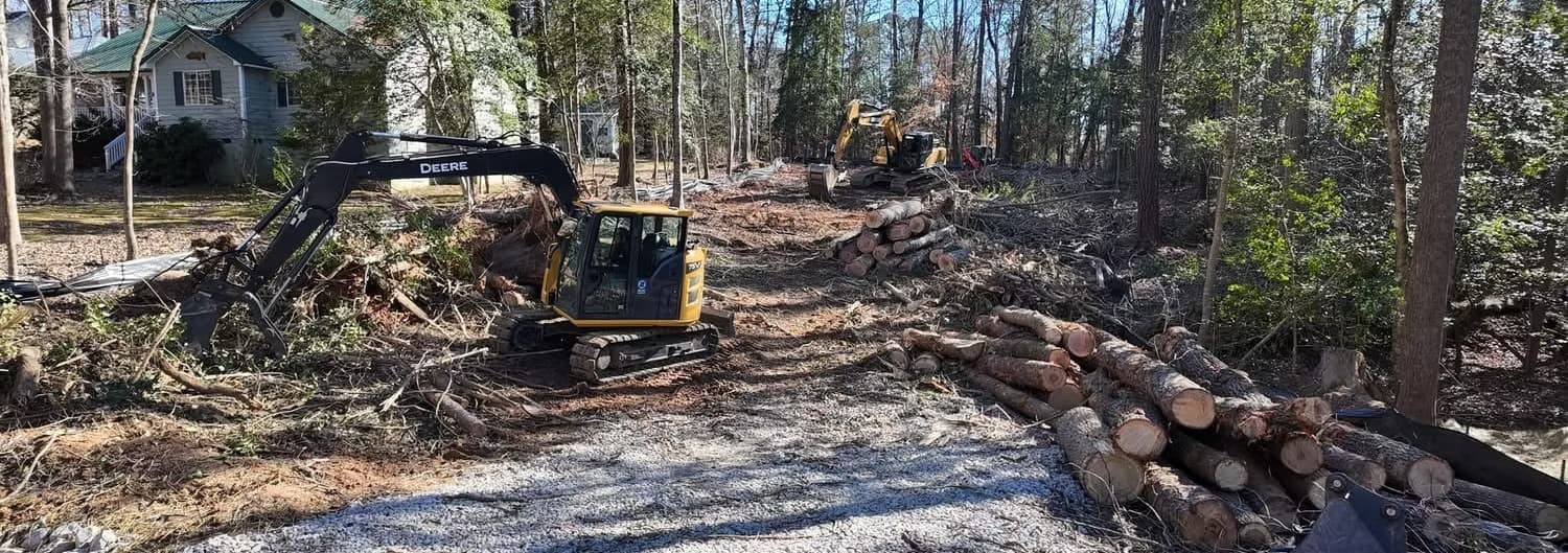An excavator clearing a wooded residential lot, with cut logs stacked along the cleared path and houses behind the tree line.