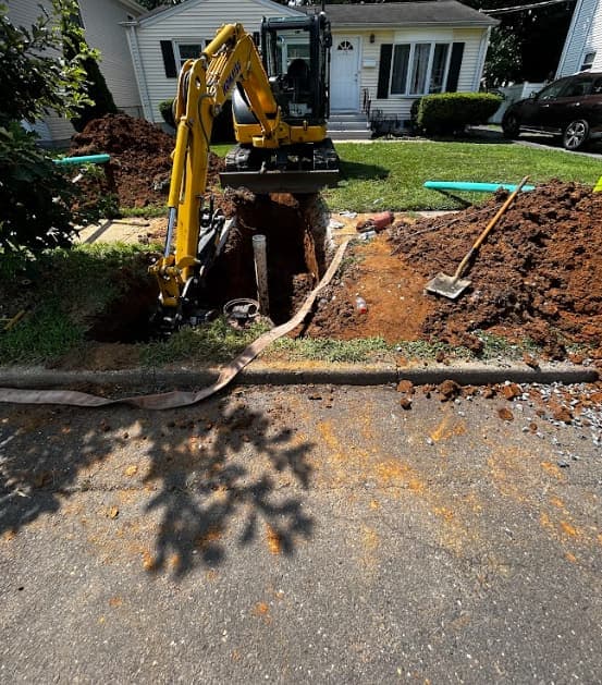 A mini excavator digging in a small front yard between the lawn and the street, spoil piled beside the hole.