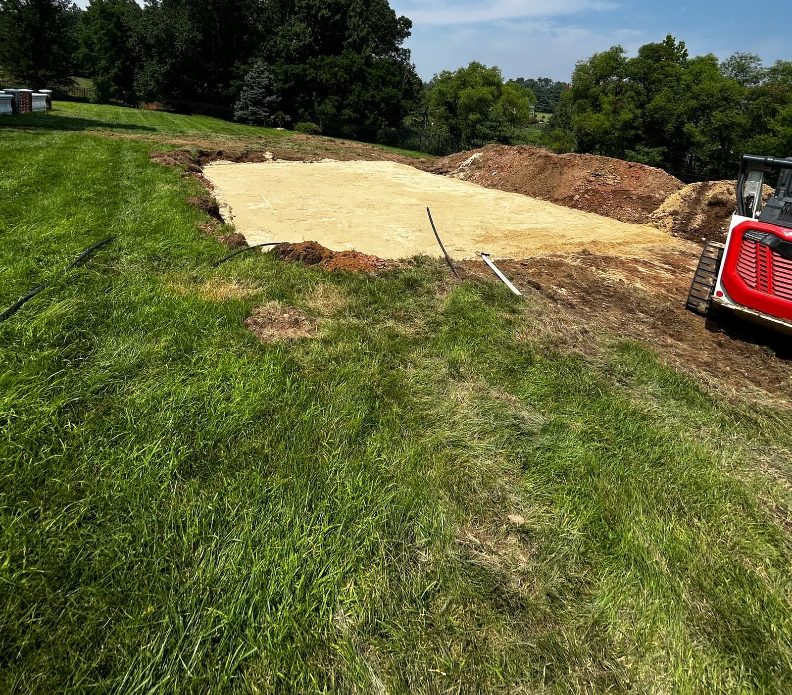 A leveled stone pad on a sloped lawn, with a tracked skid steer at the edge.