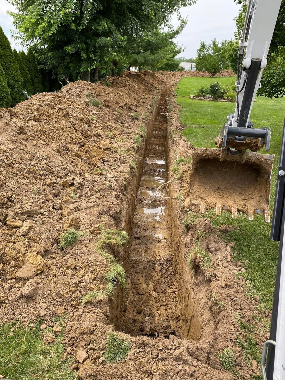 A narrow utility trench cut across a residential lawn, with the excavator bucket beside it.